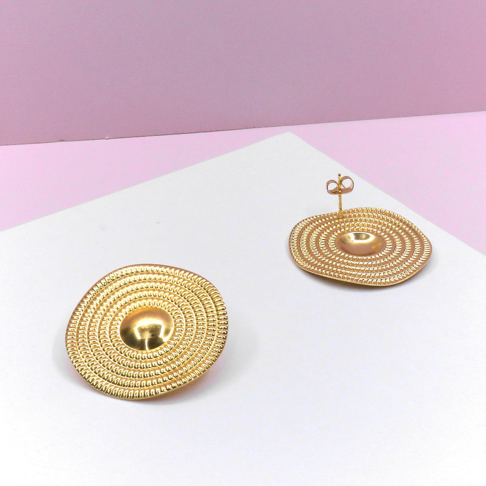Gold circular sheet earrings with textured concentric design on white and pink background