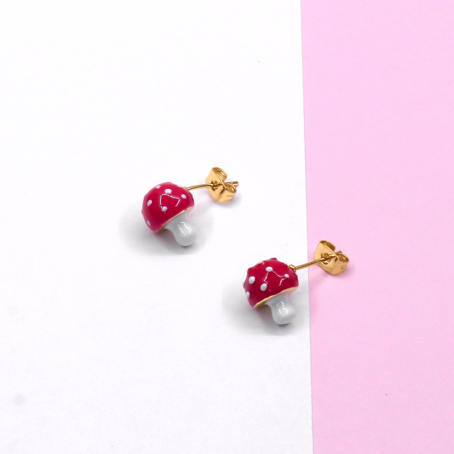 Pair of red mushroom stud earrings with white spots and gold-tone posts on white and pink background