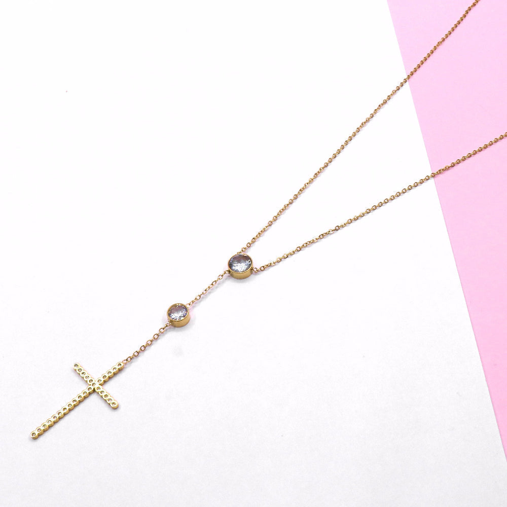 Gold cross chain necklace with two small round gemstones on a delicate chain against a white and pink background
