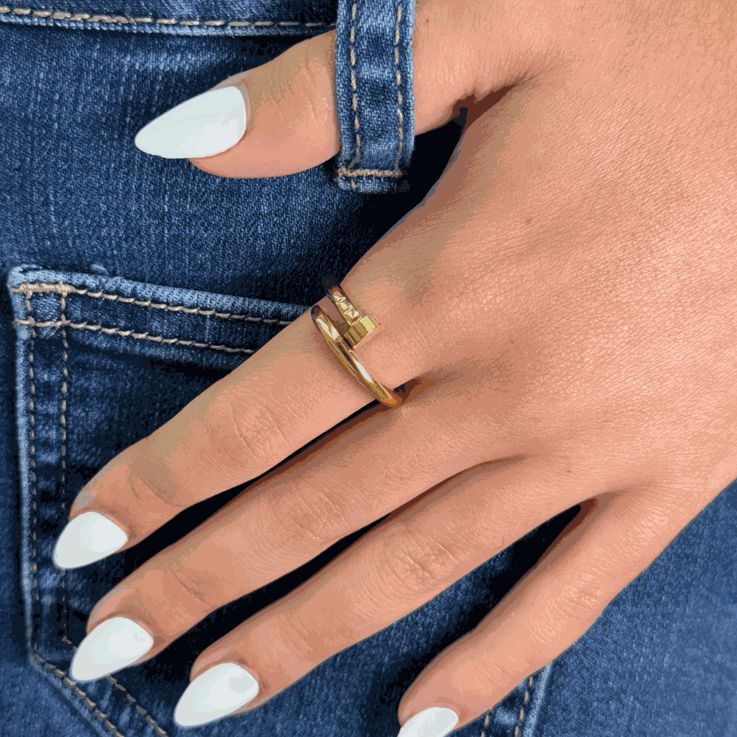 Close-up of a hand wearing a stylish gold Nail Gold Ring with manicured white nails and blue denim background