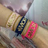 Three AMOR bracelets in pink, blue, and bright pink fabric with gold lettering and decorative gold beads on a wrist.