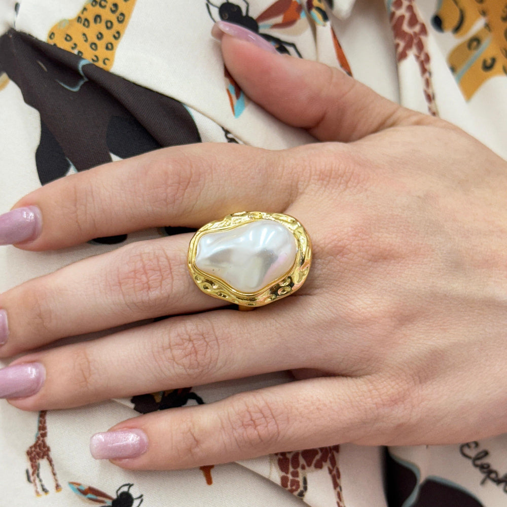 Gold ring with a heart-shaped pearl on a hand with pink nail polish, against a patterned fabric background.