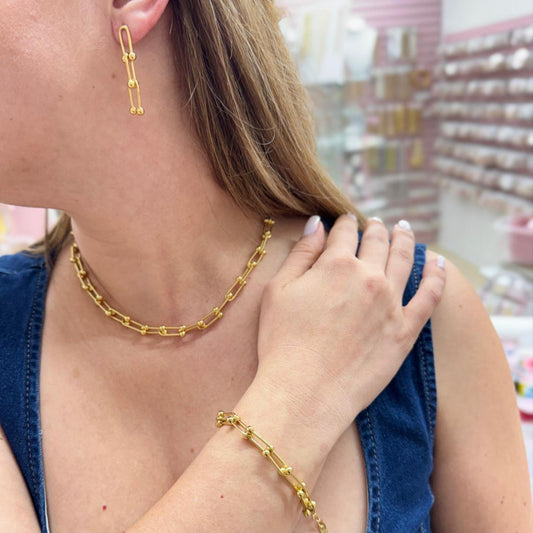 Woman wearing gold jewelry including a necklace, earrings, and bracelet in a store setting.