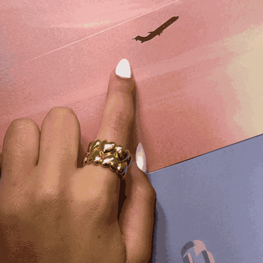 adjustable gold ring with intricate heart-shaped design worn on finger with white nail polish on pink and blue background