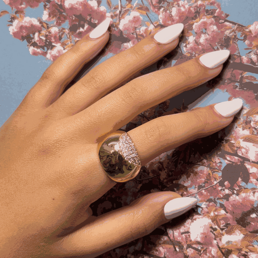 Oval gold ring with mirror-like surface and pave crystals on hand with white manicured nails against floral background