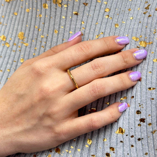 Delicate fine gold ring on hand with purple nail polish over gray fabric with gold flakes