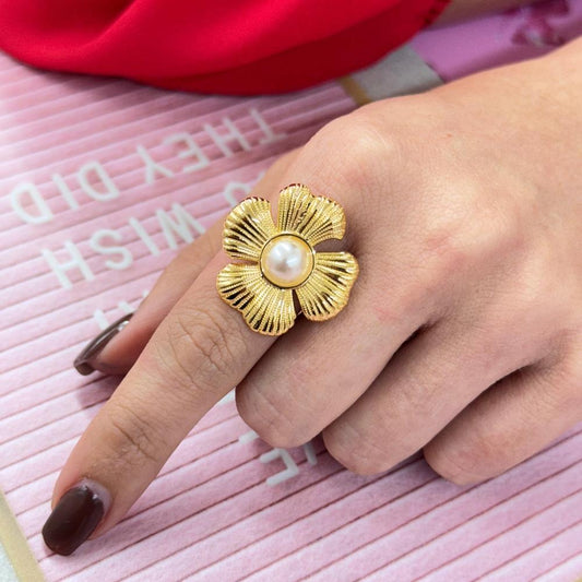 Gold flower-shaped ring with a central pearl worn on a hand with dark nail polish