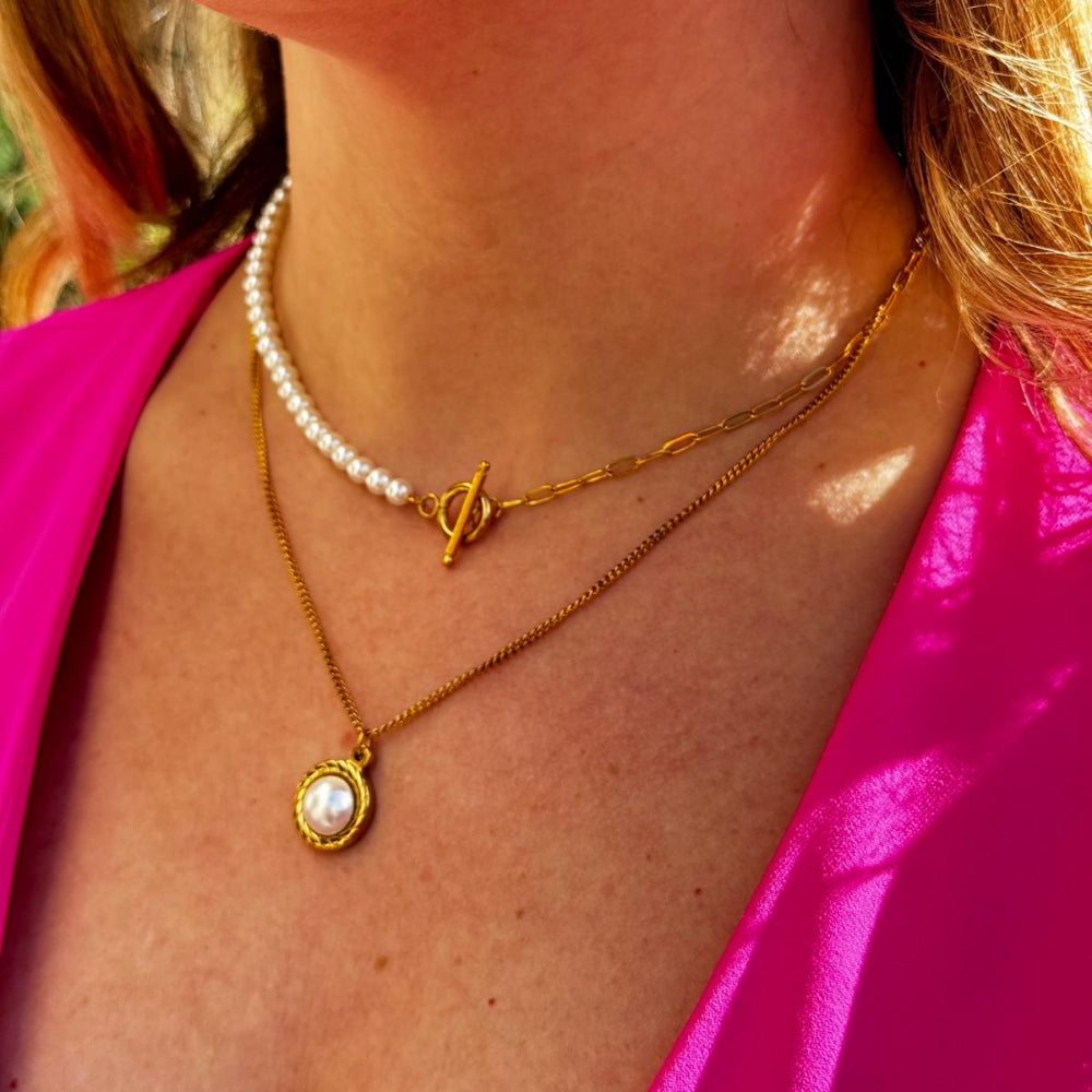 Close-up of a woman's neck wearing a luxurious pearls layering chain with gold clasp and a gold pendant necklace, styled with a bright pink top.