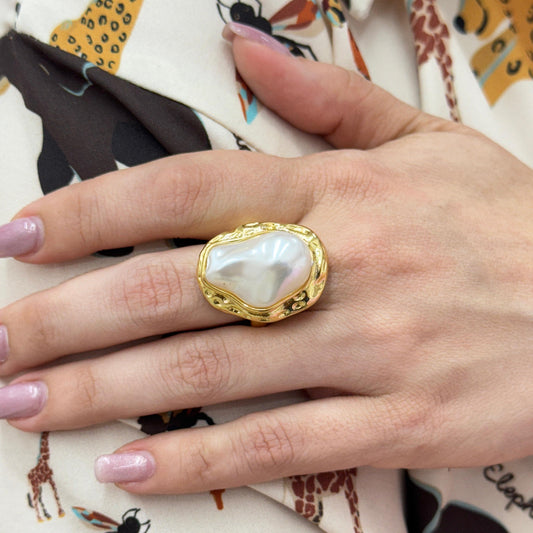 Gold ring with a heart-shaped pearl on a hand with pink nail polish, against a patterned fabric background.